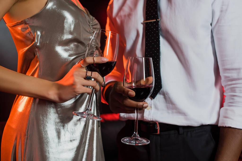 vista parcial de la mujer afroamericana y el hombre sosteniendo vasos con vino tinto en negro  - Foto, imagen
