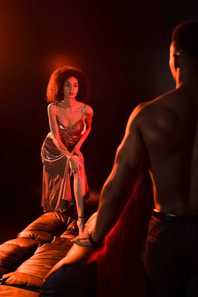 κομψή Αφρικάνικη Αμερικανίδα με φόρεμα που κοιτάζει τον γυμνό άντρα σε θολή πρόσοψη - Φωτογραφία, εικόνα