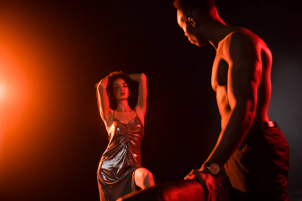 σαγηνευτική Αφροαμερικανή με φόρεμα που κοιτάζει τον γυμνό άντρα σε θολή πρόσοψη. - Φωτογραφία, εικόνα