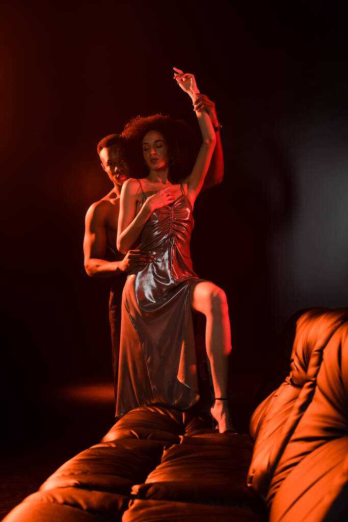 Африканский мужчина без рубашки соблазняет женщину в платье возле дивана на черном  - Фото, изображение