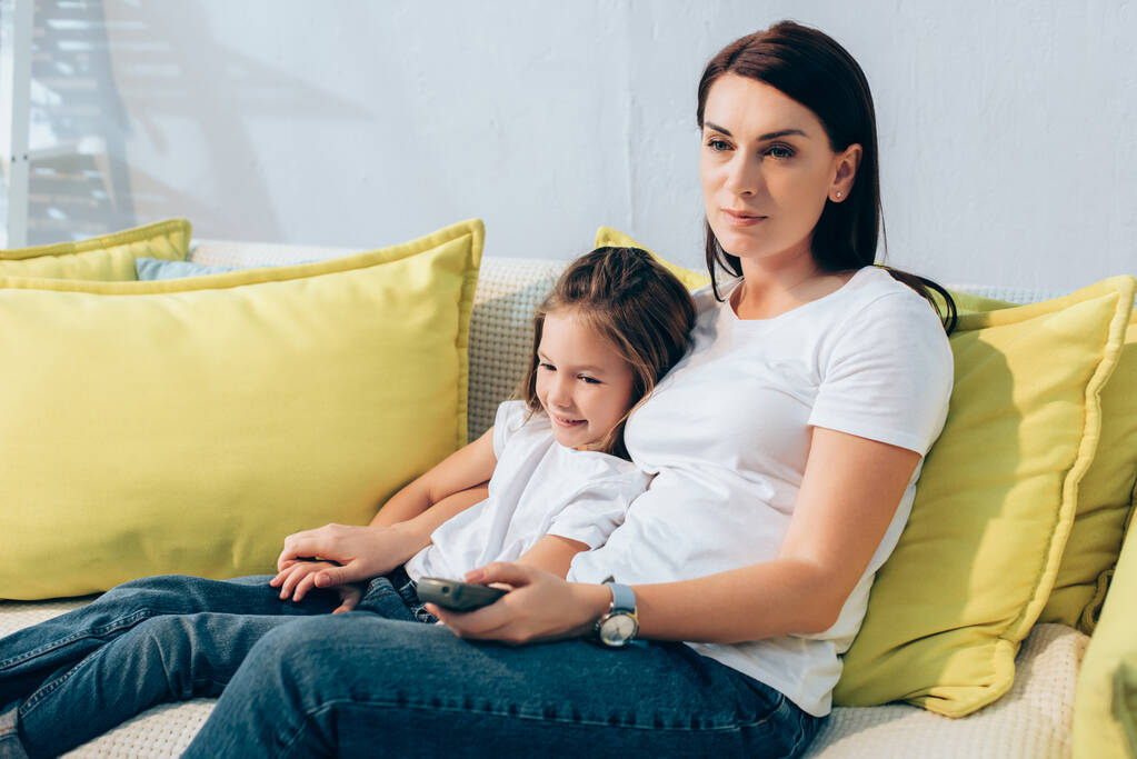 Madre con telecomando abbracciare sorridente figlia mentre seduto sul divano - Foto, immagini