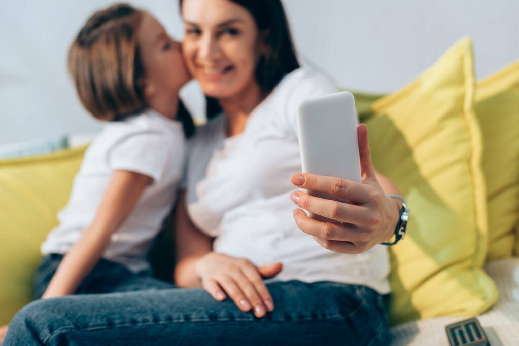 Hija besando sonriente madre durante selfie en casa en borrosa fondo - Foto, imagen