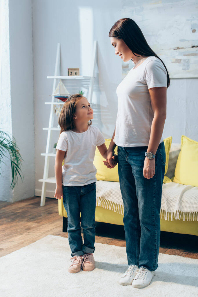 Full length of smiling mother and daughter looking at each other while holding hands at home on blurred background - Photo, Image