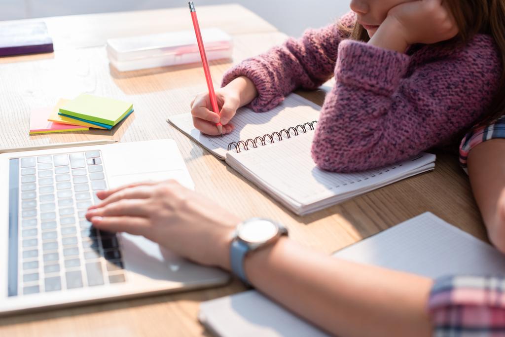 Gewassen weergave van dochter schrijven in notebook aan het bureau met wazig moeder typen op laptop op de voorgrond - Foto, afbeelding