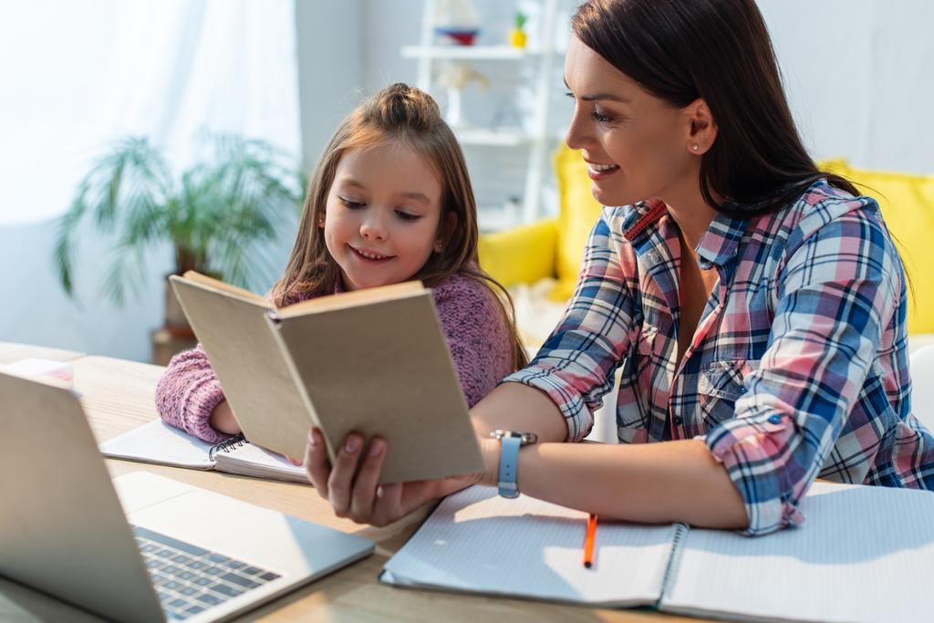 Улыбающиеся мать и дочь читают книгу за столом с ноутбуком на размытом фоне - Фото, изображение