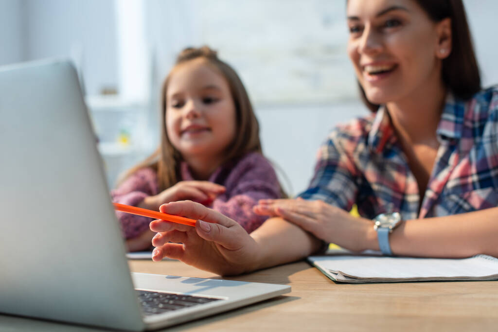 Uśmiechnięta matka wskazująca ołówkiem na laptopa w pobliżu córki w domu na rozmazanym tle - Zdjęcie, obraz