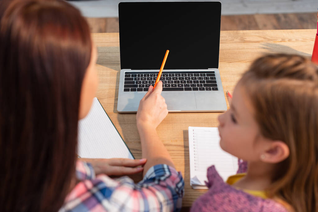 Vue grand angle de la mère pointant avec un crayon à l'ordinateur portable près de la fille sur le premier plan flou - Photo, image