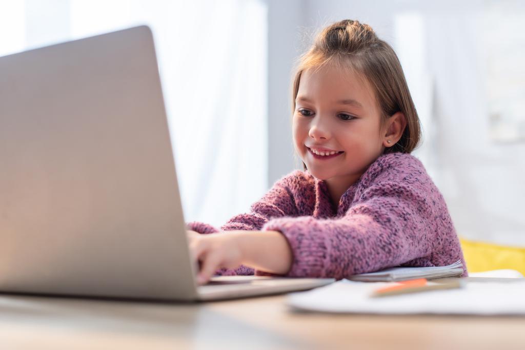 Χαμογελαστή δακτυλογράφηση κορίτσι στο laptop στο γραφείο σε θολή πρώτο πλάνο - Φωτογραφία, εικόνα