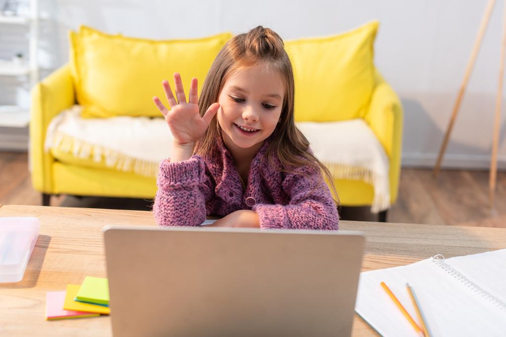 Lächelndes Mädchen mit winkender Hand und Blick auf Laptop auf Schreibtisch mit Papeterie im verschwommenen Vordergrund - Foto, Bild