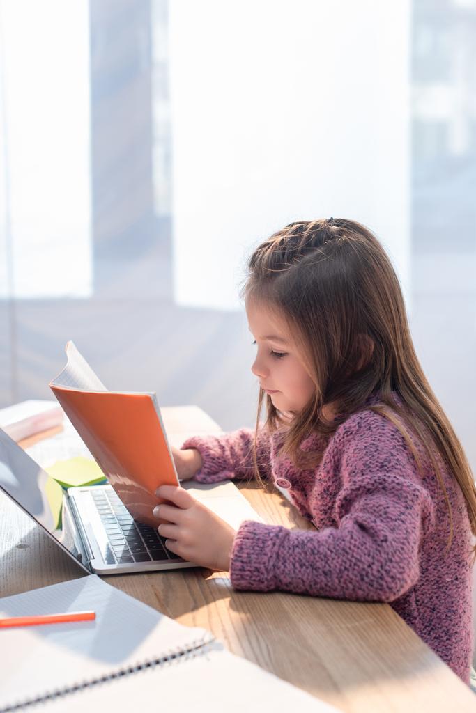 Mädchen blickt auf Kopierbuch neben Laptop auf Schreibtisch im verschwommenen Vordergrund - Foto, Bild