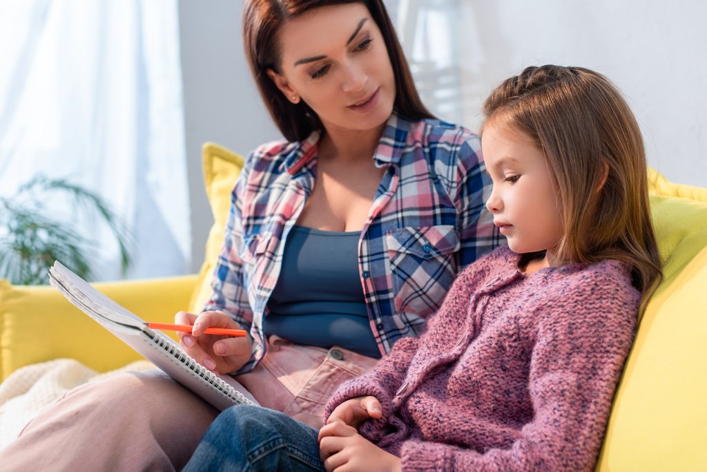 Madre con matita e copiare libro guardando la figlia sul divano su sfondo sfocato - Foto, immagini