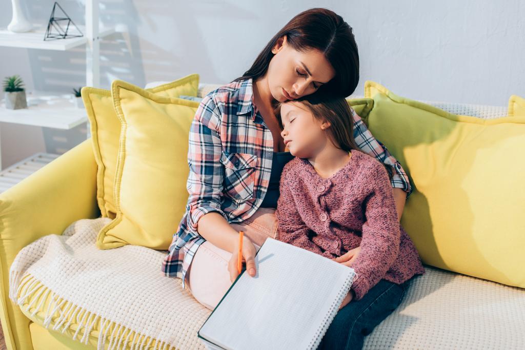 Madre molesta con lápiz y libro de copias abrazando a la hija durmiendo en el sofá - Foto, Imagen