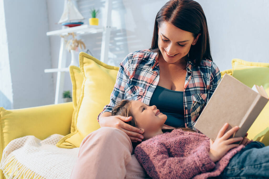 Lächelnde Mutter blickt Tochter mit Buch auf Sofa auf verschwommenem Hintergrund an - Foto, Bild