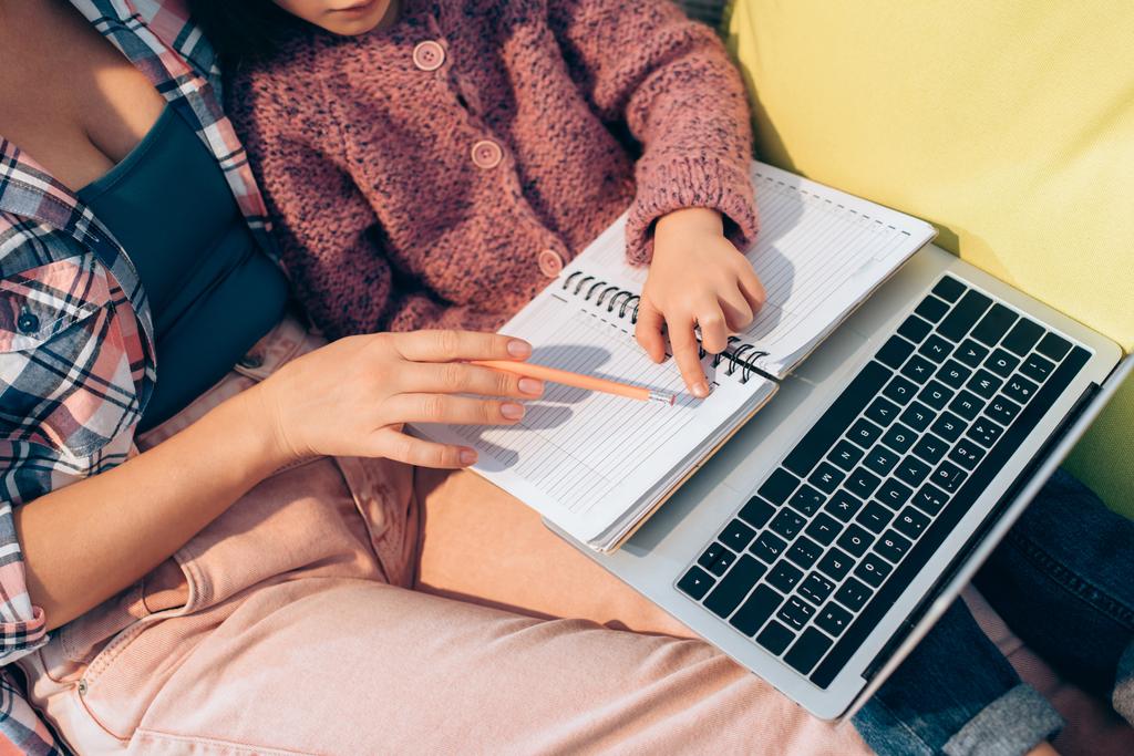 Обрезанный вид дочери указывая пальцем на ноутбук рядом с матерью с карандашом и ноутбуком на диване - Фото, изображение