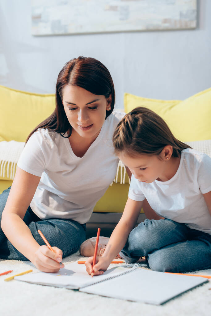 Улыбающиеся мать и дочь рисуют цветными карандашами на полу на размытом фоне - Фото, изображение