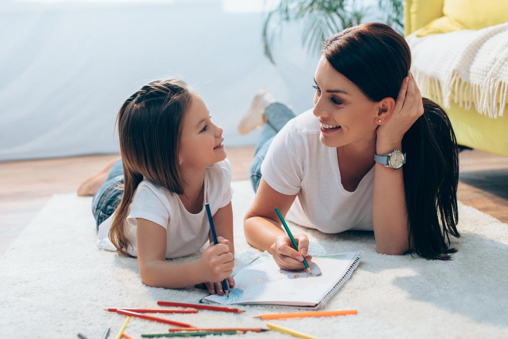 Cheerful mother and daughter with colored pencils looking at each other while lying on floor on blurred background - Photo, Image