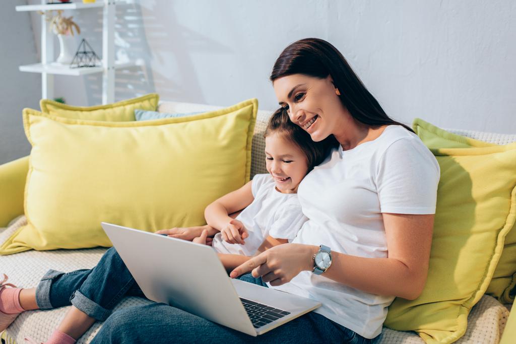 Щаслива мати і дочка сміються, вказуючи пальцями на ноутбук на дивані на розмитому фоні
 - Фото, зображення