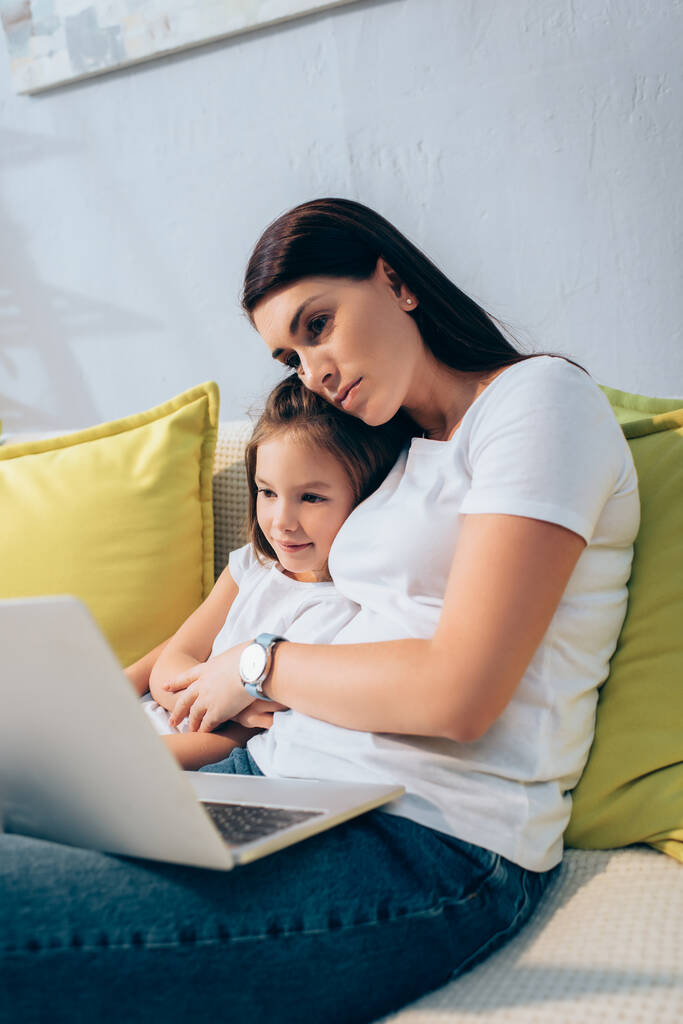 Mama przytula uśmiechniętą córkę patrząc na laptopa na kanapie na niewyraźnym pierwszym planie - Zdjęcie, obraz