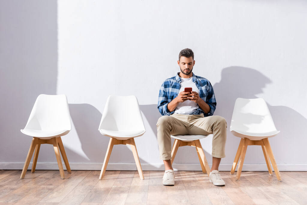 Homme en vêtements décontractés en utilisant smartphone tout en attendant sur la chaise dans le hall  - Photo, image