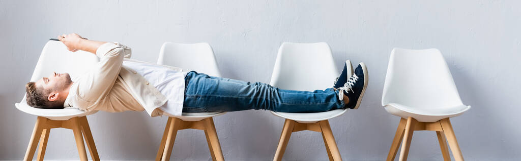 Homme allongé sur des chaises et utilisant un smartphone en attendant dans le hall, bannière  - Photo, image