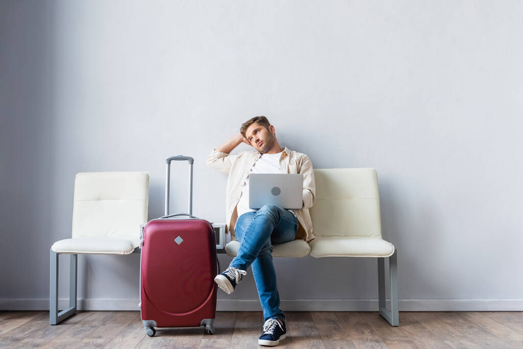 Uomo con laptop che guarda lontano vicino alla valigia mentre aspetta in aeroporto  - Foto, immagini