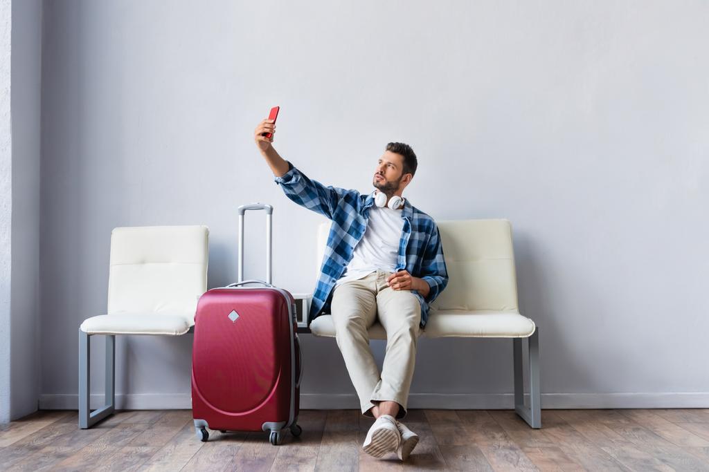 Muž ve sluchátkách se selfie s chytrým telefonem v blízkosti kufru na letišti  - Fotografie, Obrázek