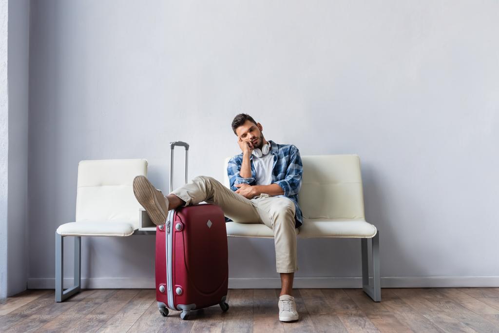 Müder Mann mit Kopfhörern sitzt neben Koffer, während er in Flughafenhalle wartet  - Foto, Bild