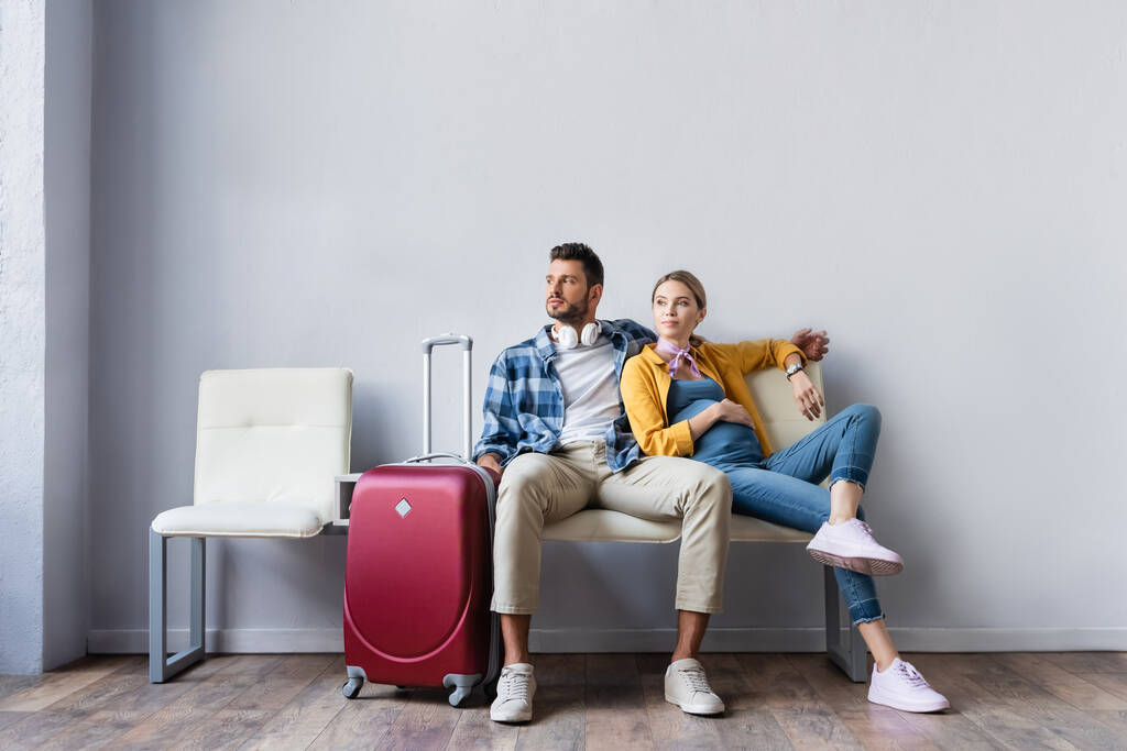 Вагітна жінка сидить біля чоловіка і валізи в аеропорту
  - Фото, зображення