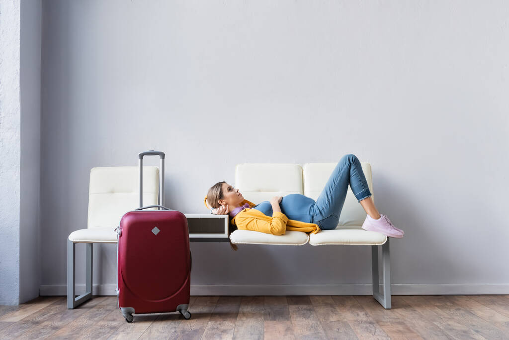 Ciężarna kobieta leżąca na krzesłach w pobliżu walizki w holu lotniska  - Zdjęcie, obraz