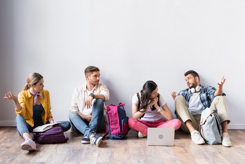 Мультикультурные студенты с рюкзаками ждут экзамен возле рюкзаков и ноутбука на полу  - Фото, изображение