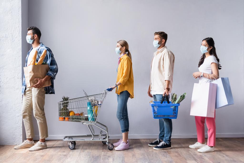Gente multicultural en máscaras médicas sosteniendo carrito de la compra, cesta con comida y bolsas en cola en el mercado  - Foto, imagen