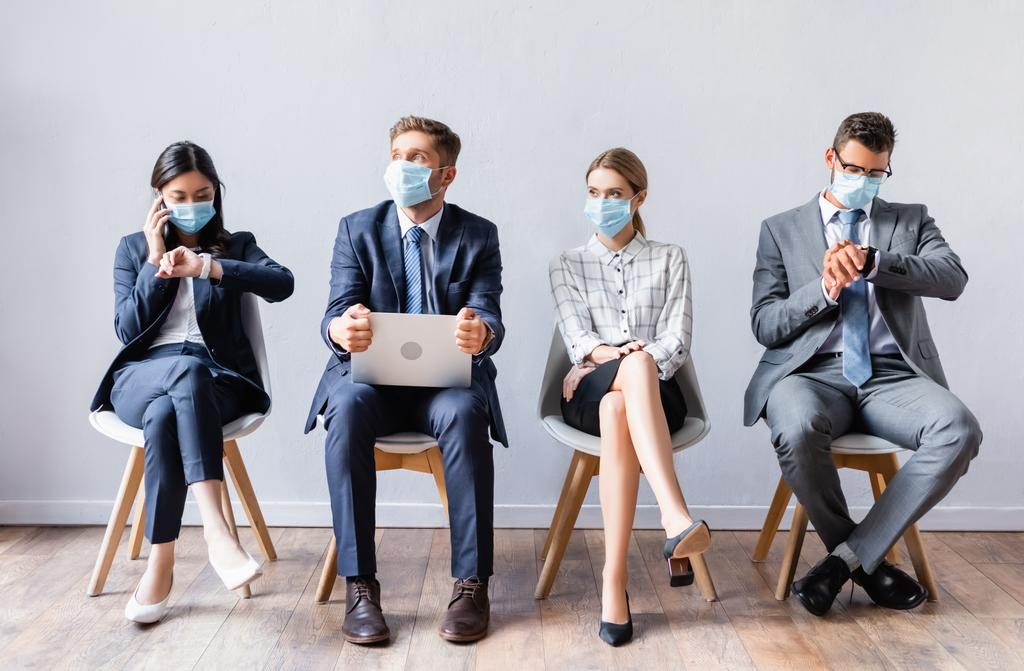 Multikulturelle Menschen in medizinischen Masken mit Laptop und Smartphone warten auf Stühlen in der Halle  - Foto, Bild