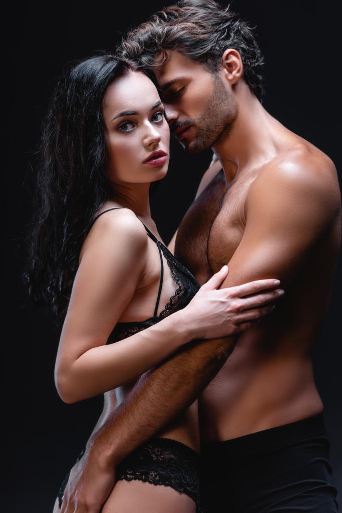 сексуальный мужчина без рубашки обнимает чувственную брюнетку женщина смотрит на камеру изолированы на черный - Фото, изображение