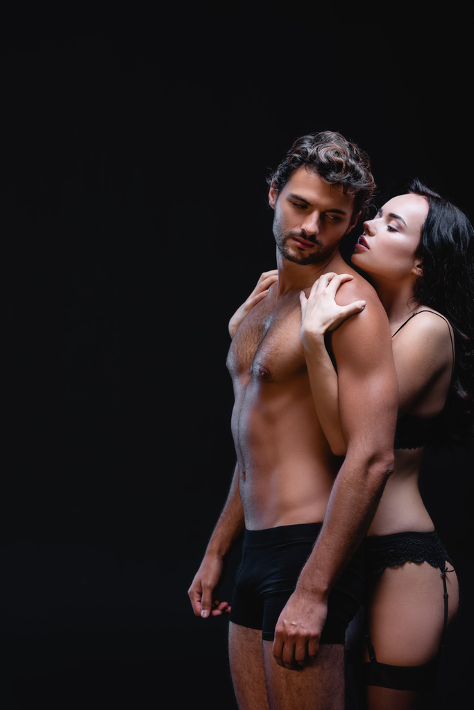 svůdná žena objímající ramena sexy mladý muž izolované na černé - Fotografie, Obrázek