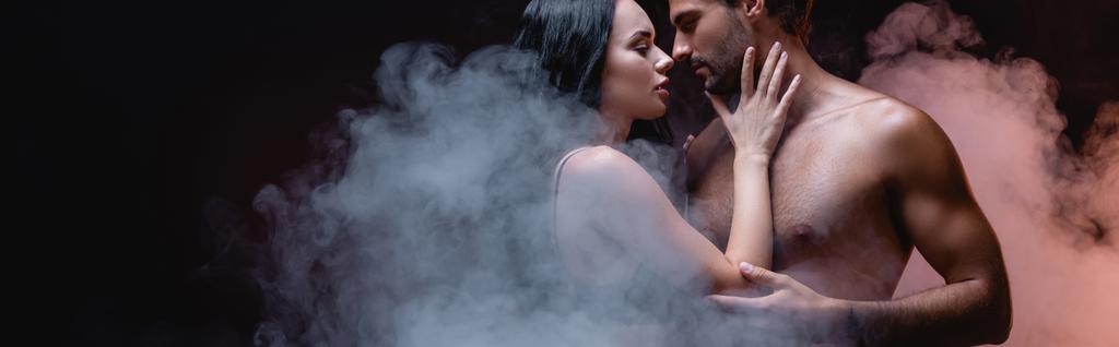 сексуальная женщина прикасается к мужчине без рубашки стоя лицом к лицу на черном фоне с дымом, баннер - Фото, изображение