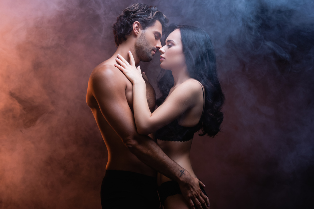 Мужчина без рубашки обнимает соблазнительную женщину, стоя лицом к лицу на темном фоне с дымом - Фото, изображение