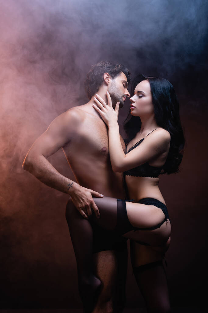 femme sexy en lingerie noire et bas touchant le cou de l'homme torse nu sur fond sombre avec de la fumée - Photo, image