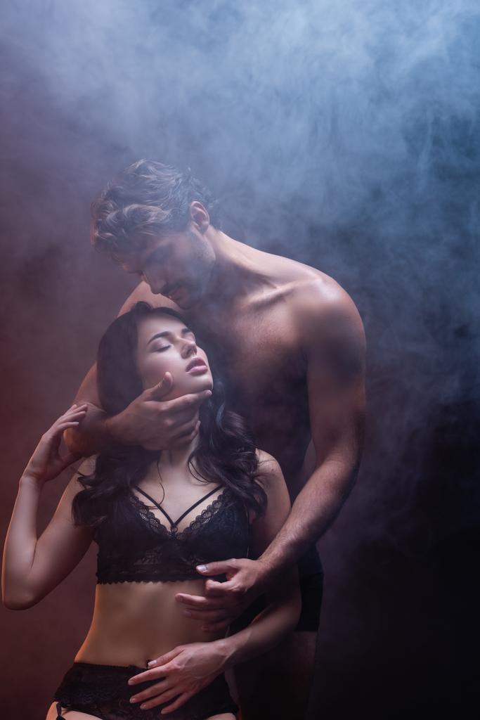 sexy hemdloser Mann umarmt Hals der verführerischen Frau in schwarzen Spitzenunterwäsche auf dunklem Hintergrund mit Rauch - Foto, Bild