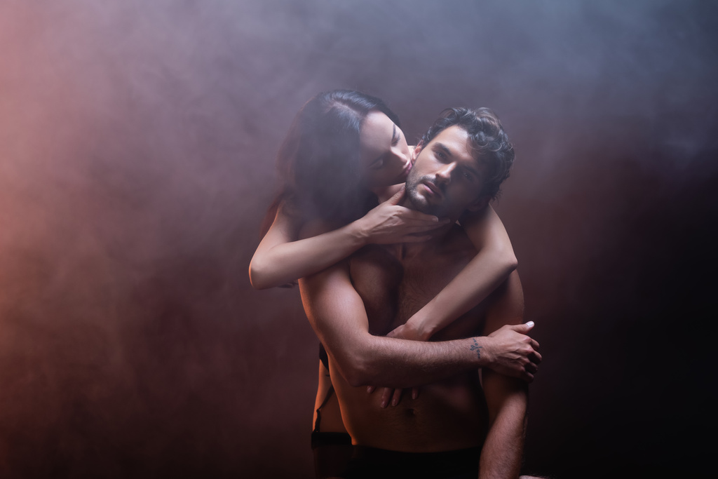 sensual mulher abraçando e beijando jovem sem camisa homem olhando para câmera no fundo escuro com fumaça - Foto, Imagem