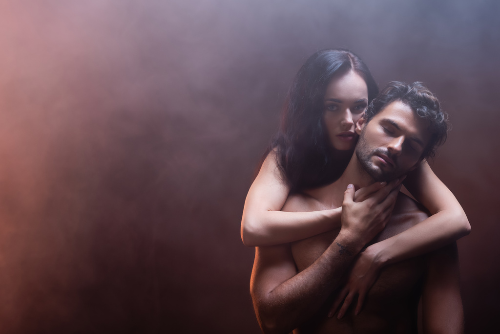 mujer seductora mirando a la cámara mientras abraza al hombre sin camisa sobre fondo oscuro con humo - Foto, Imagen