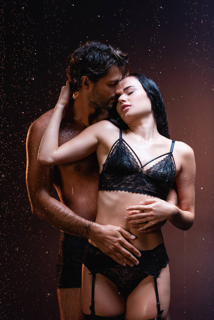 hombre joven abrazando a mujer seductora en lencería de encaje negro bajo la lluvia sobre fondo oscuro - Foto, Imagen