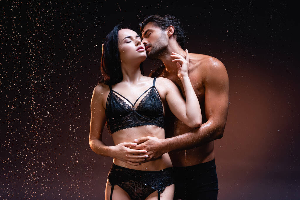 hemdloser Mann umarmt und küsst sexy Frau in schwarzer Unterwäsche unter strömendem Regen auf dunklem Hintergrund - Foto, Bild