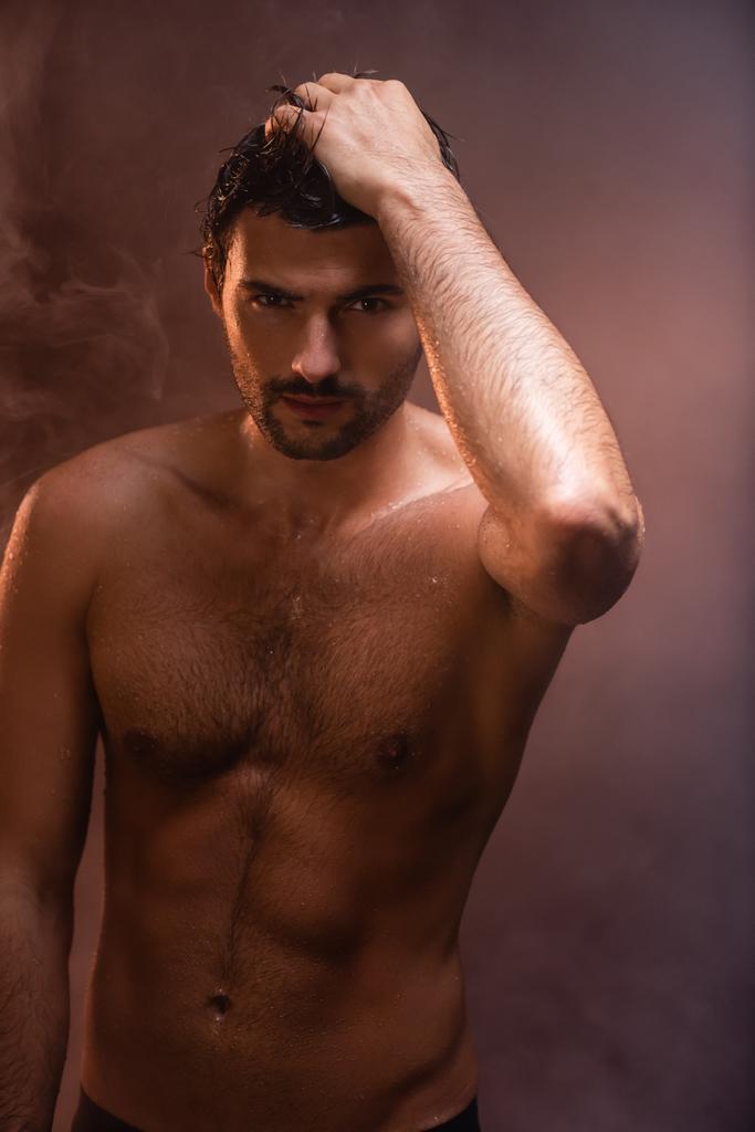 セクシーな濡れた男で筋肉質の胴は煙で暗い背景の上に手でポーズをとって - 写真・画像