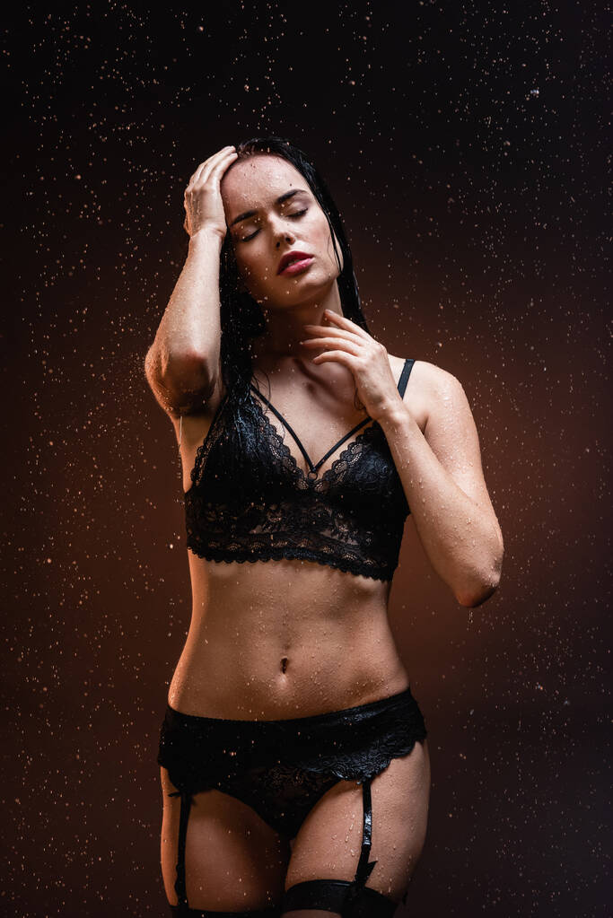 mujer sexy en ropa interior de encaje negro y medias con los ojos cerrados bajo gotas de lluvia cayendo sobre fondo oscuro - Foto, Imagen