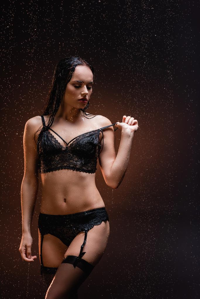 mujer joven en ropa interior de encaje negro tocando tira de sujetador bajo lluvia cayendo sobre fondo oscuro - Foto, imagen