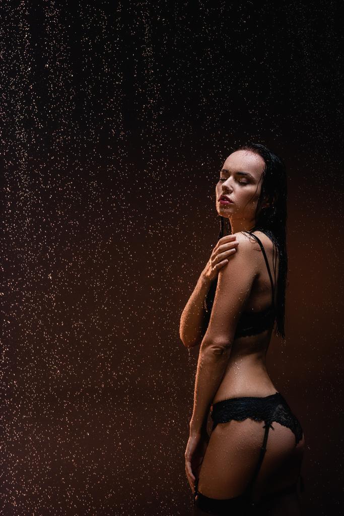 verleidelijke vrouw in zwart kant lingerie met gesloten ogen onder regen op donkere achtergrond - Foto, afbeelding