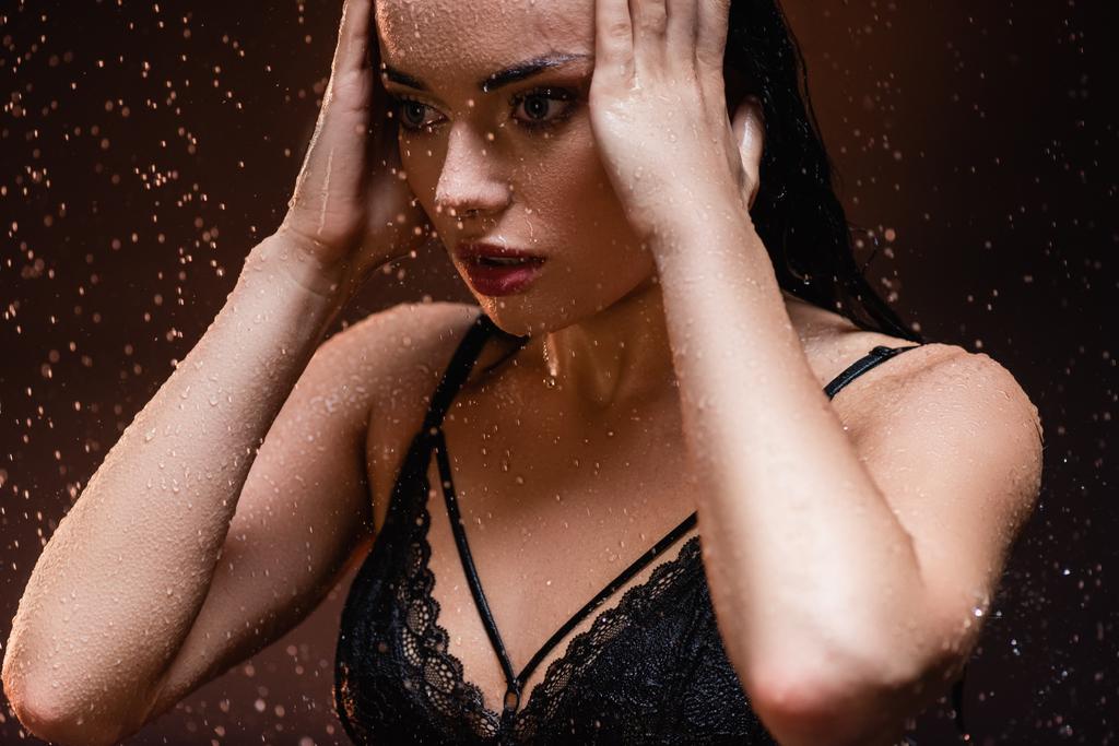 mujer sensual en sujetador de encaje negro posando con las manos cerca de la cara bajo la lluvia sobre fondo oscuro - Foto, imagen