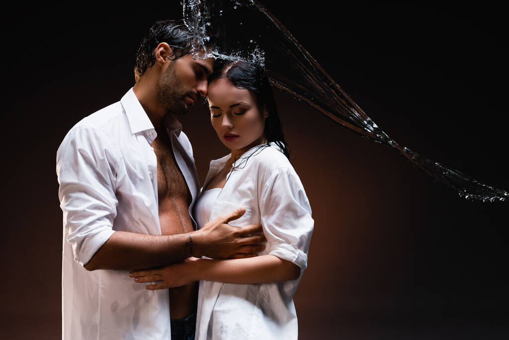 junges Paar in weißen nassen Hemden umarmt sich mit geschlossenen Augen in der Nähe von Wasserspritzern auf dunklem Hintergrund - Foto, Bild