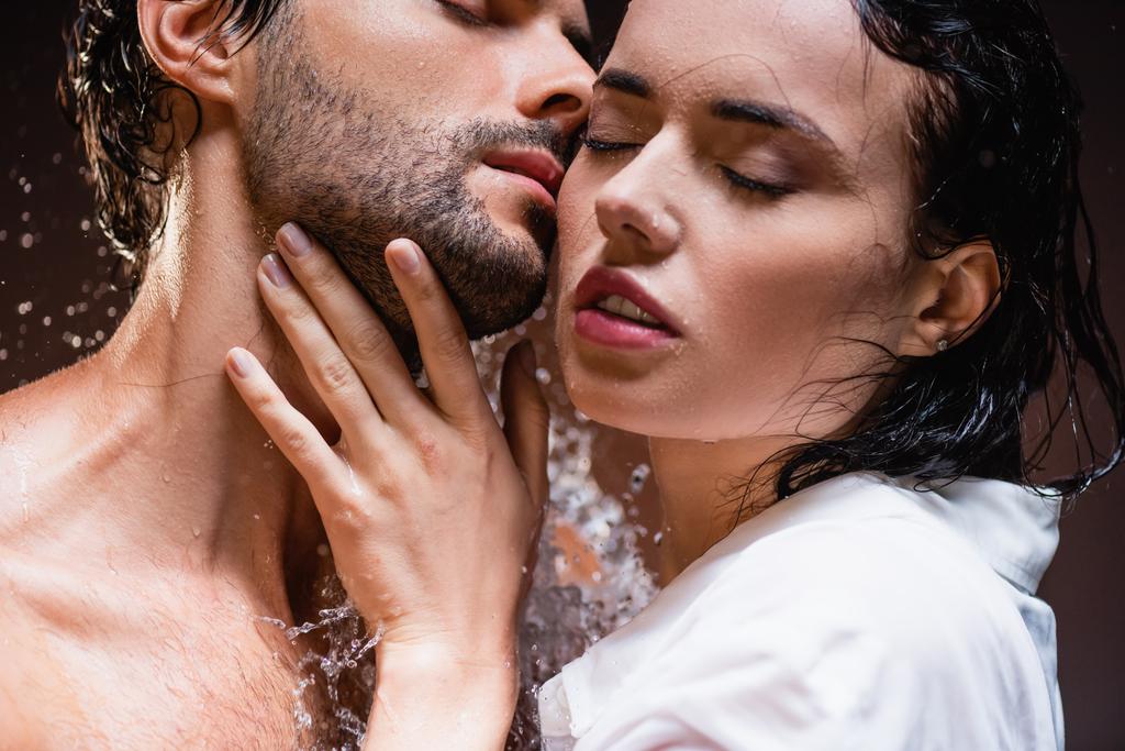 donna seducente toccare giovane uomo con gli occhi chiusi vicino spruzzi d'acqua su sfondo scuro - Foto, immagini