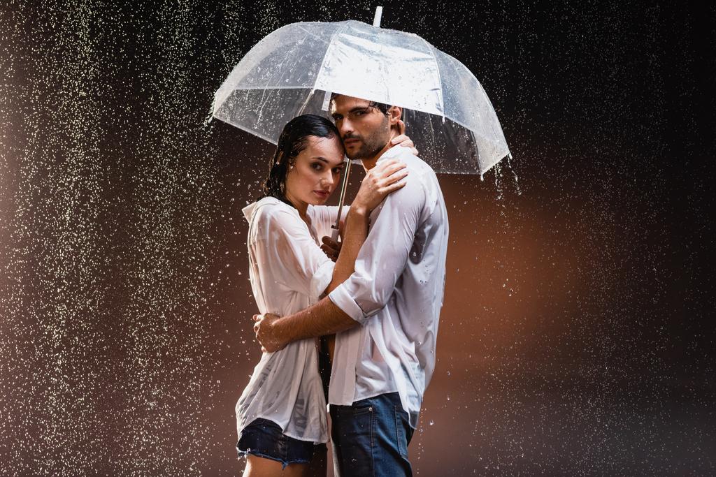 young couple in wet shirts standing under rain with transparent umbrella on dark background - Photo, Image